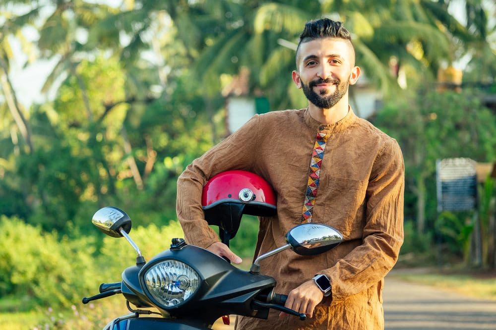 A smiling man standing by his scooter with a helmet under his arm